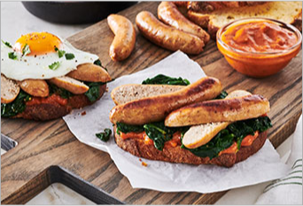 two toasts topped with sausages, spinach, egg and sauce on a wooden board