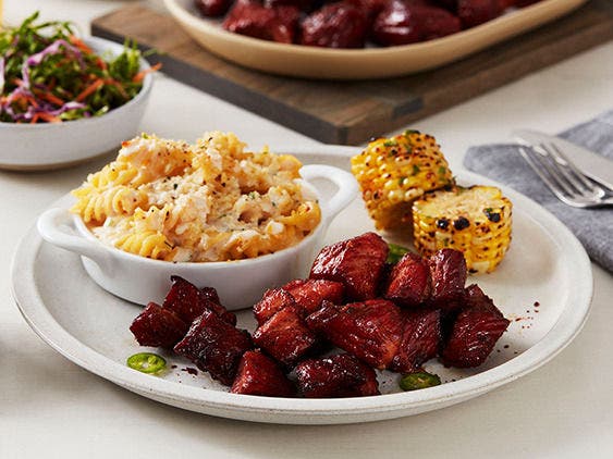 Pork shoulder burnt ends on a plate with grilled corn on the cob and pasta salad.