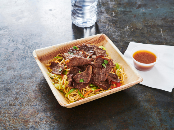Philly beef on noodles with veggies, served on a wooden plate and sauce on the side