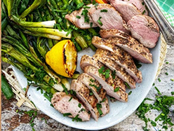 Grilled pork tenderloin cut up, next to a grilled lemon and asparagus. 