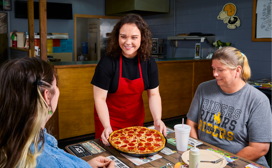 Girl on a red apron serving a pepperoni pizza to two women customers